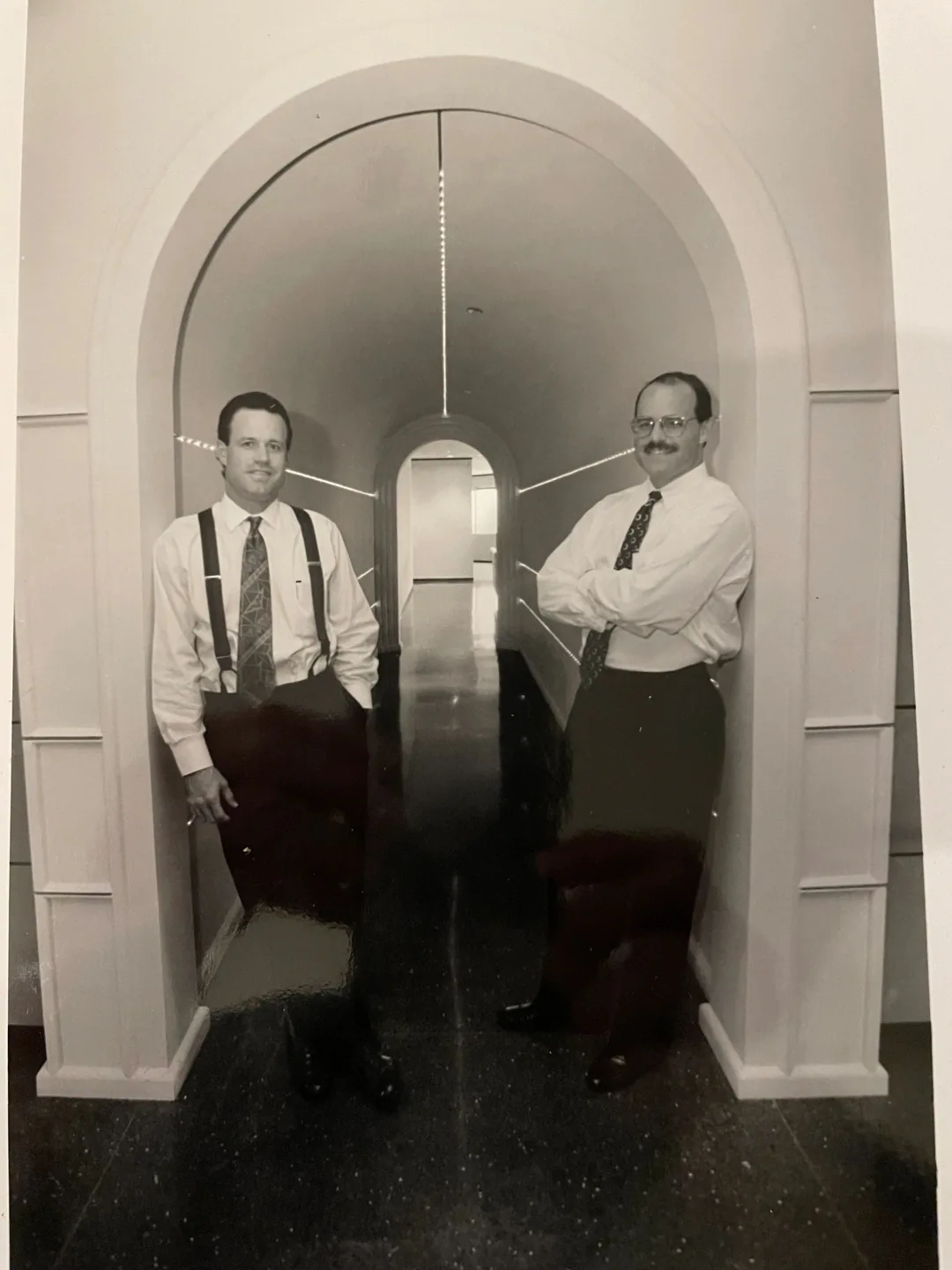 Two men in formal attire stand in an arched hallway, one leaning and the other with arms crossed.